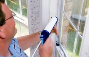 Man wearing glasses applies caulk with a caulk gun to a window frame inside a house.